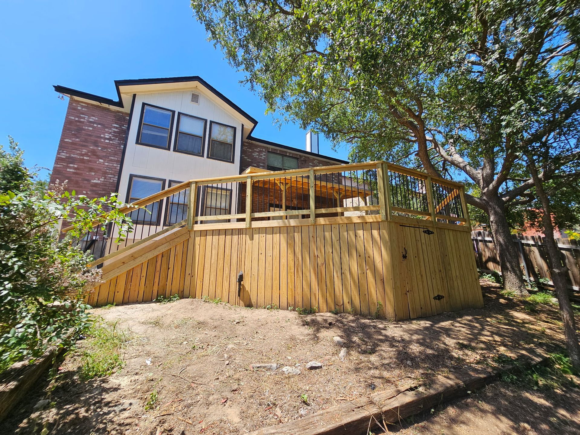 Wooden deck attached to a two-story house, surrounded by trees and grass.