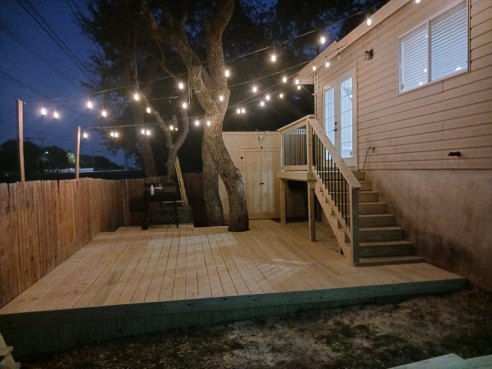Wooden deck with string lights, a grill, and stairs leading to a house at dusk. A fence and trees are also visible.