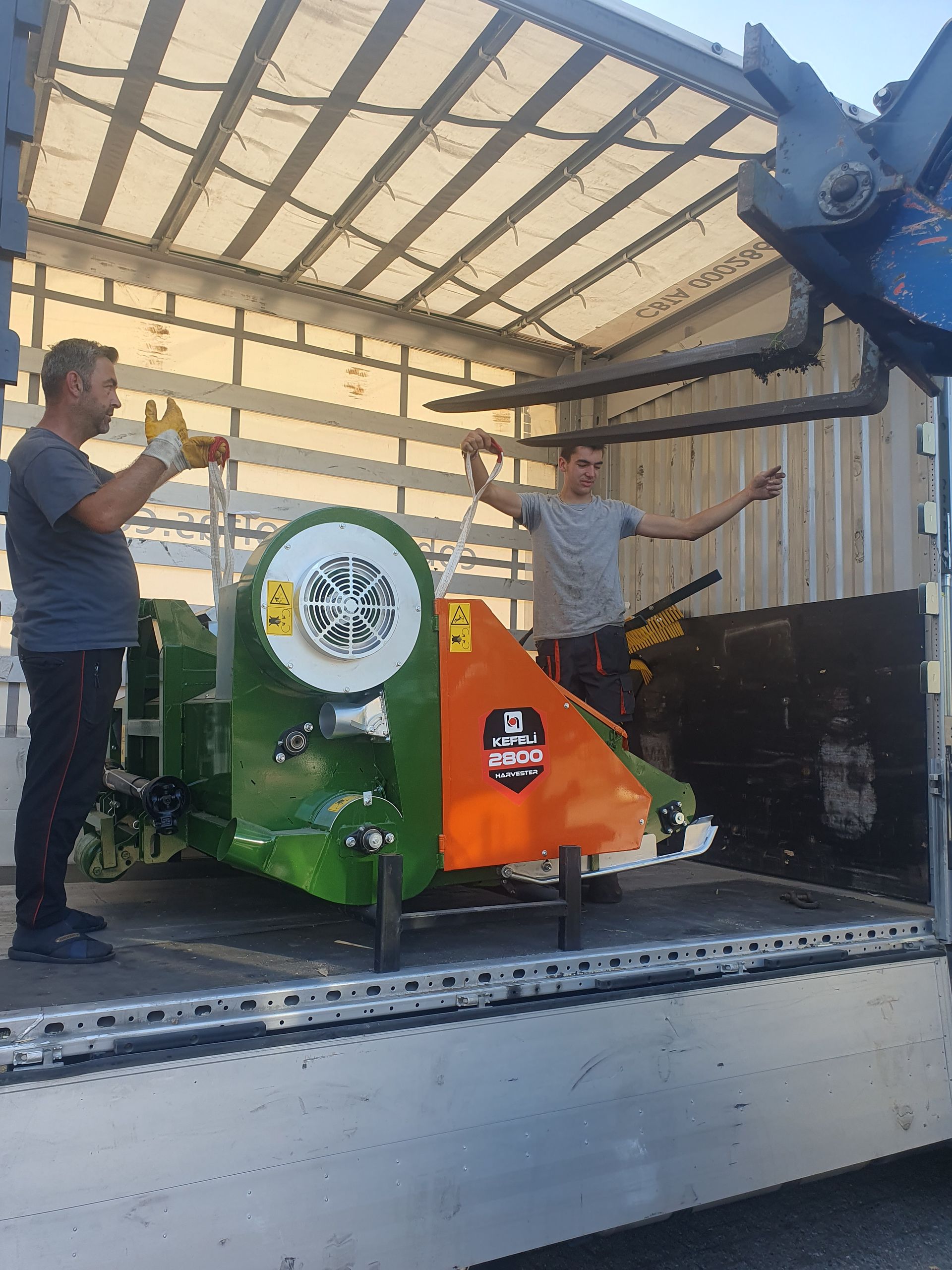 Twee mannen laden een groen-oranje machine in een vrachtwagen. Een van de mannen steekt zijn armen uit.