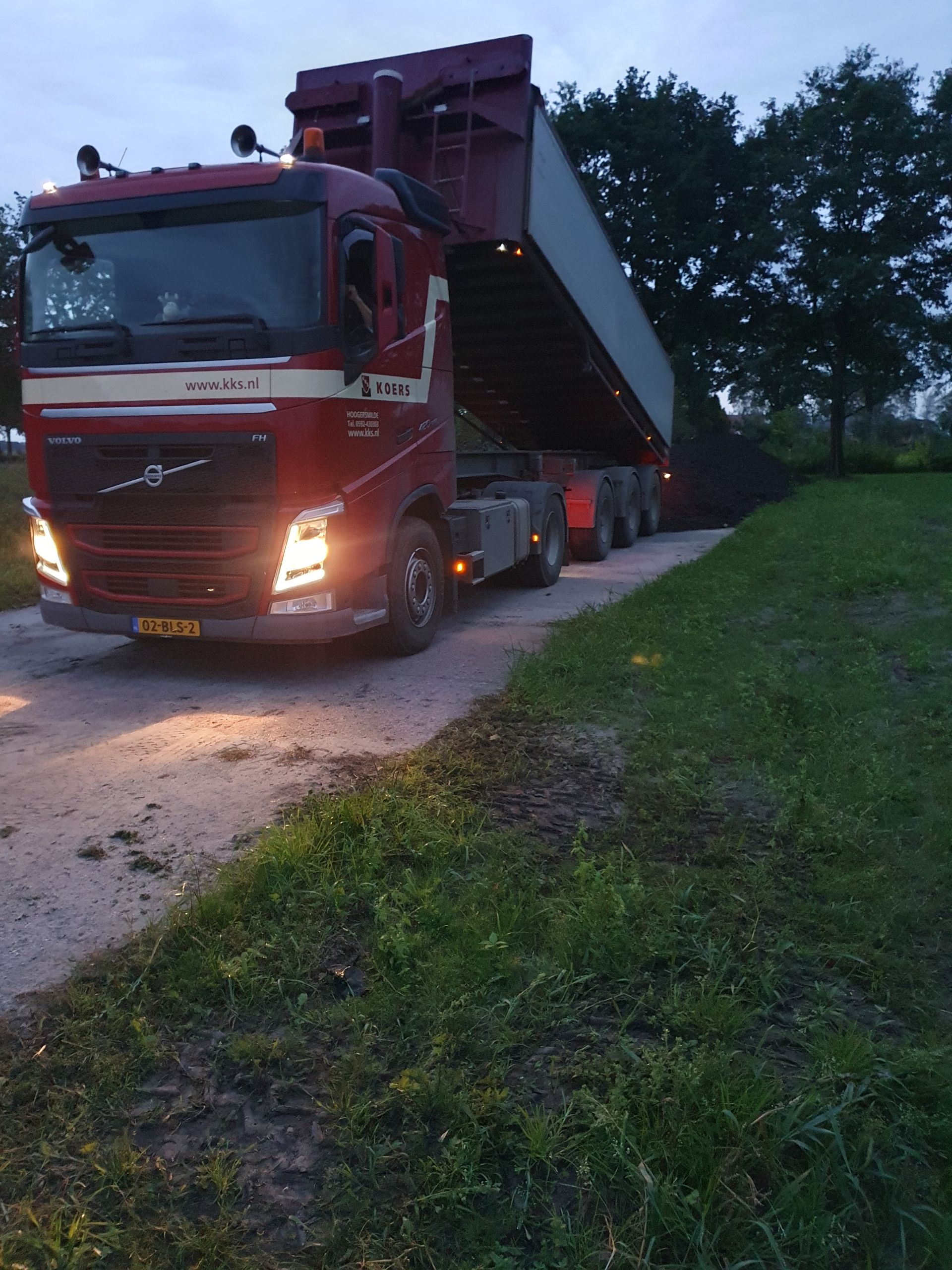 Rode Volvo-kipper die aan het lossen is op een grindpad, gras aan de rechterkant, bomen op de achtergrond.