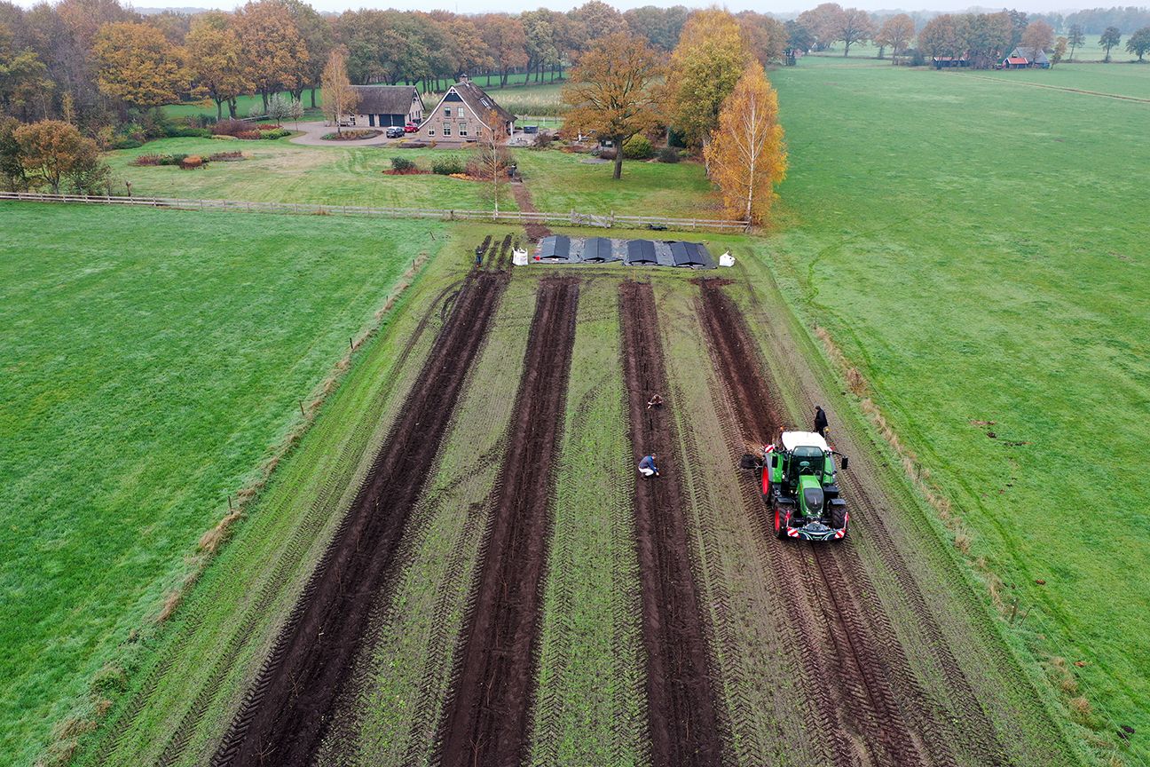 Een tractor ploegt donkere grond in lange rijen op een veld, met een boerderij en bomen op de achtergrond.
