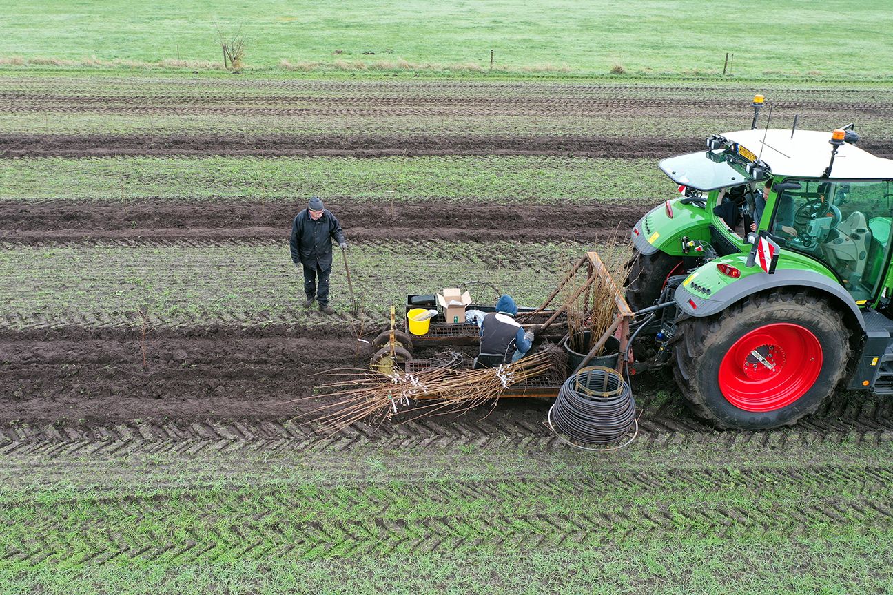 Een tractor trekt een zaaimachine door een modderig veld. Een persoon in een donkere jas staat er vlakbij.