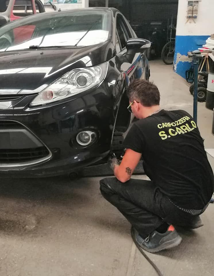 Un meccanico è accovacciato accanto al paraurti anteriore di un'auto nera in un'officina meccanica.