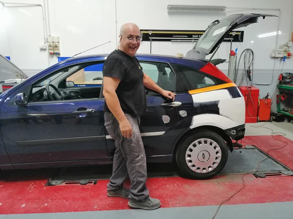 Un uomo è in piedi accanto a una berlina blu in un'officina, con il portellone posteriore aperto e i pannelli della carrozzeria a vista.