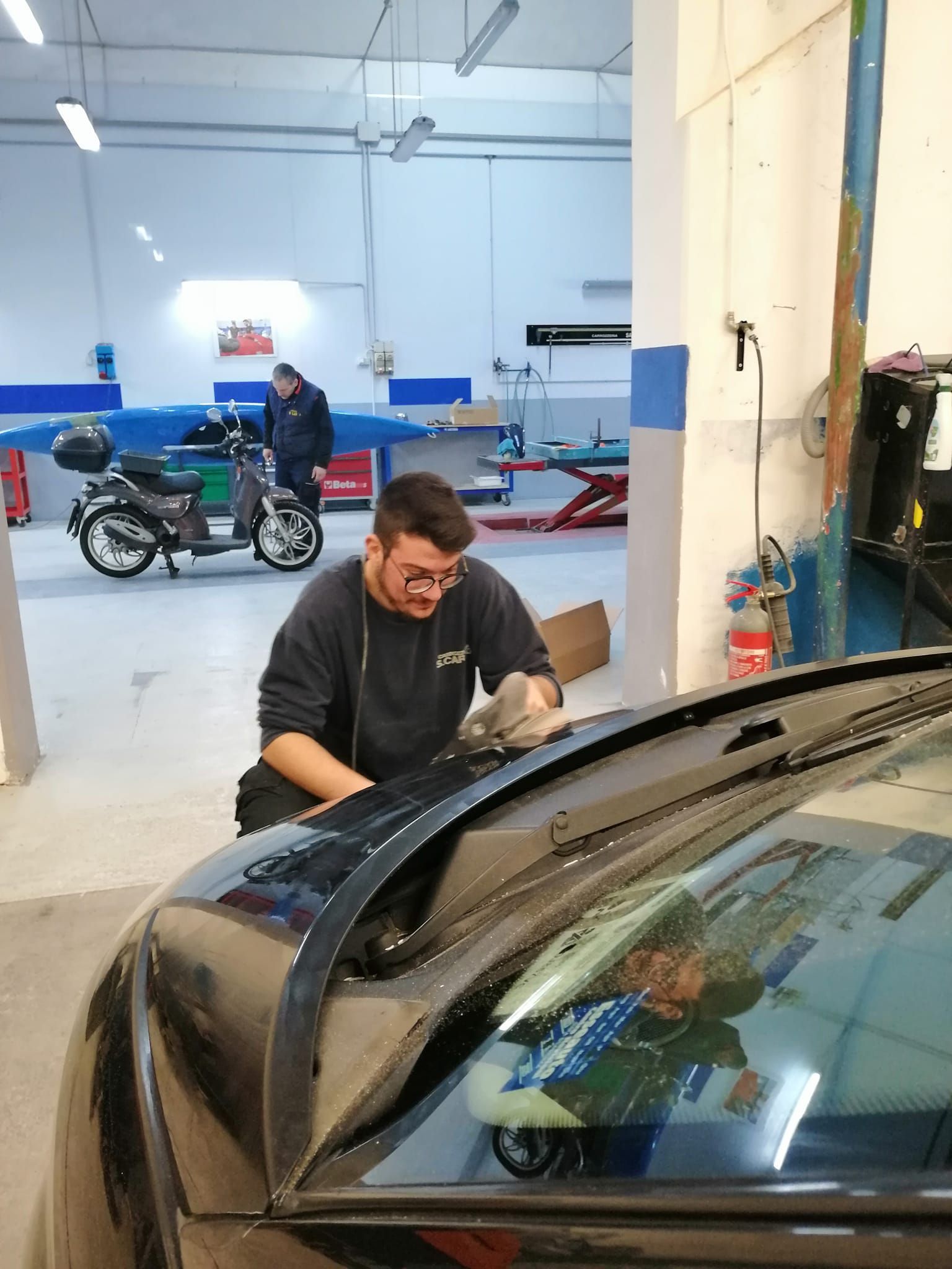 Un meccanico lucida un'auto nera in una luminosa officina, con delle motociclette sullo sfondo.