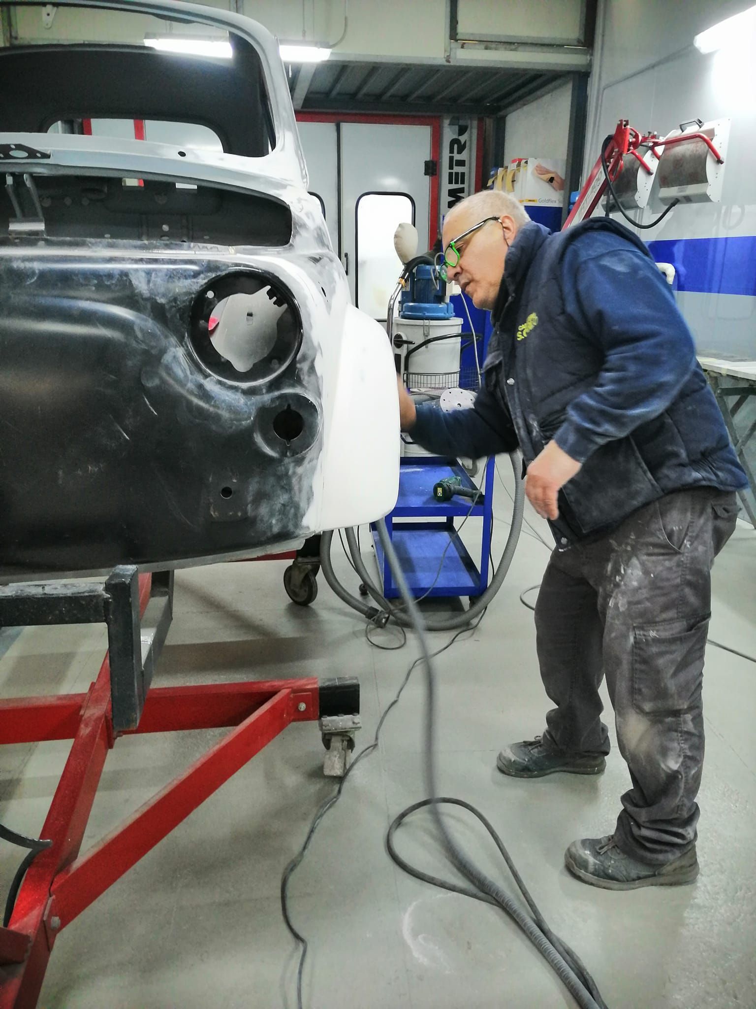 Un operaio carteggia la carrozzeria di un'auto in un'officina meccanica, con polvere di carteggiatura e attrezzi ben visibili.