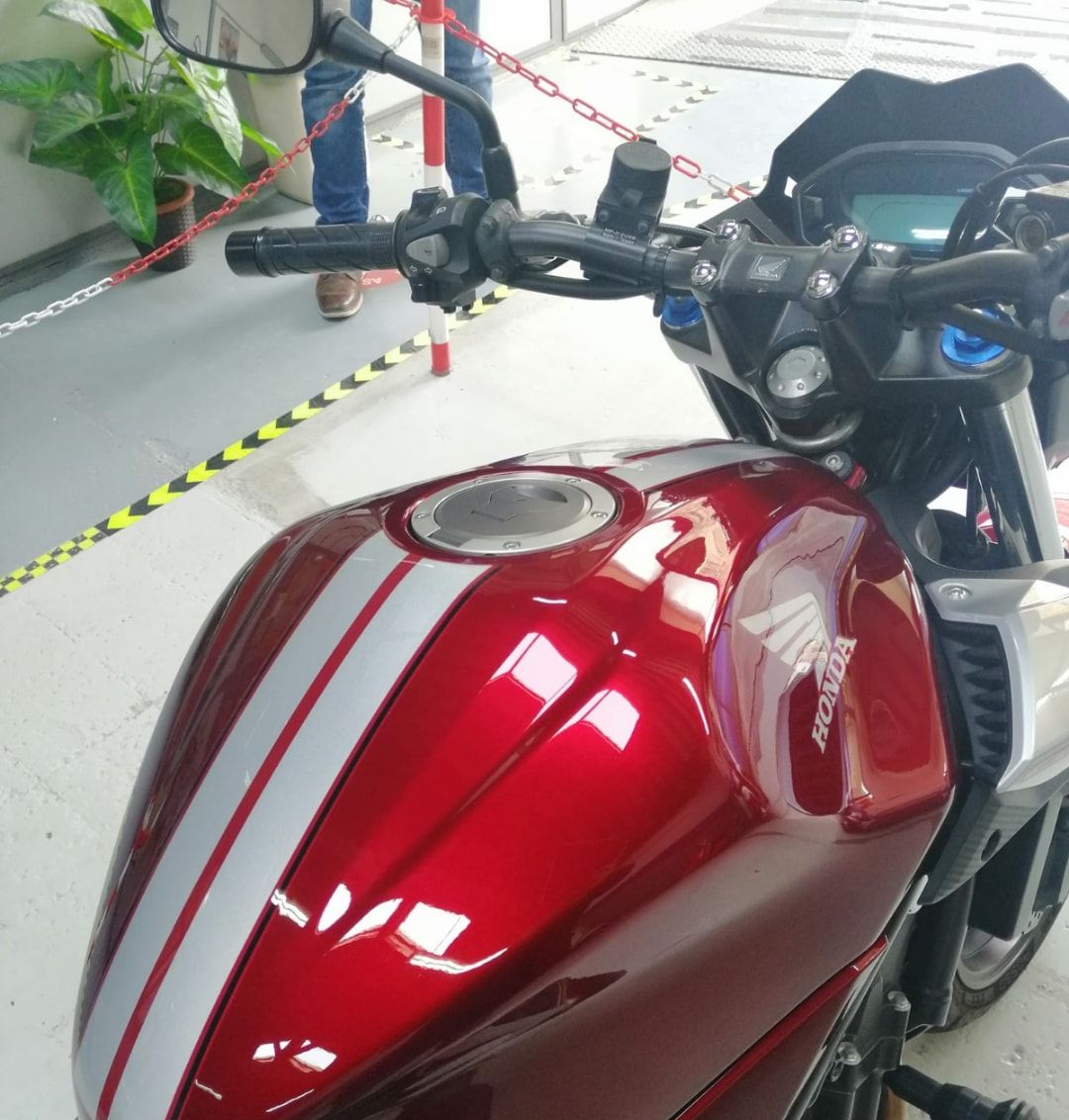 Serbatoio e manubrio di una motocicletta rossa parcheggiati all'interno di un edificio, dietro una barriera di segnalazione.