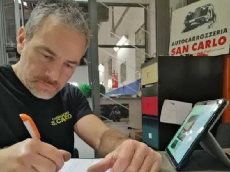 Un uomo con una camicia nera scrive su un foglio di carta a una scrivania, con un tablet e l'insegna dell'Autocarrozzeria San Carlo sullo sfondo.