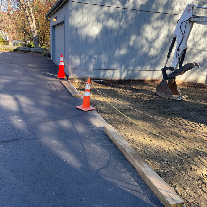 Construction site with orange cones, lumber, and an excavator next to a driveway and garage.