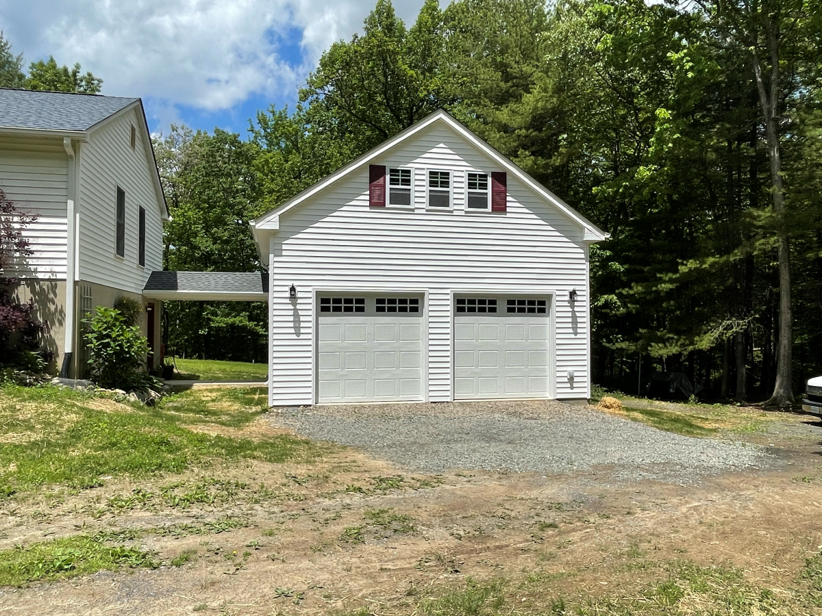 White two-car garage with red shutters, connected to a house via a covered walkway, in a wooded area.