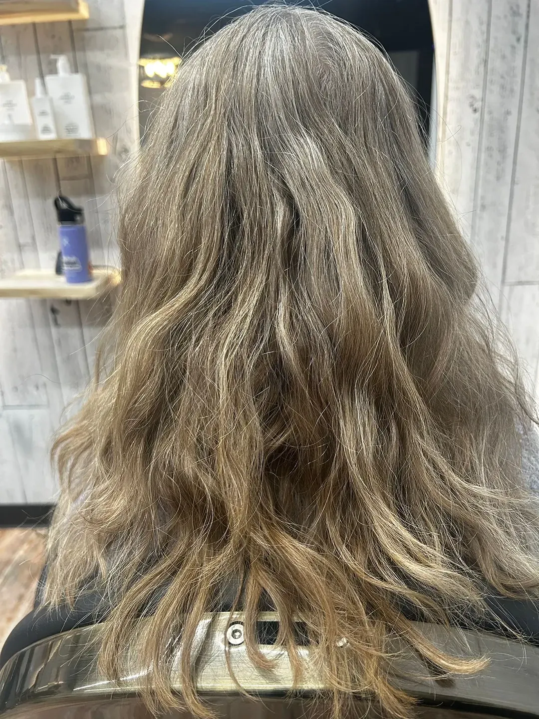 Back view of a person with wavy light-brown hair inside a salon, seated before a mirror.