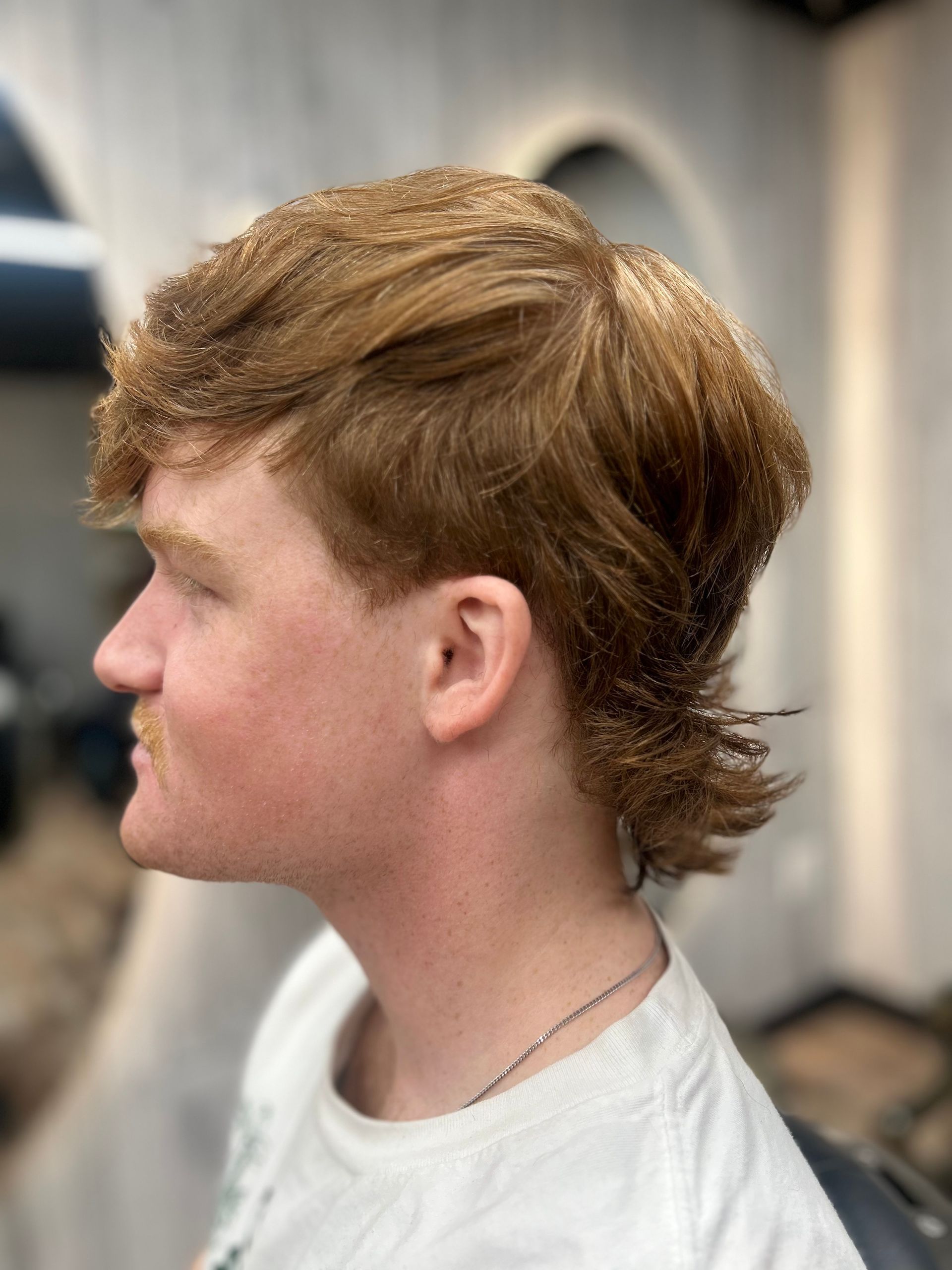 Side-profile portrait of a person with short reddish hair, wearing a white shirt, in a salon setting