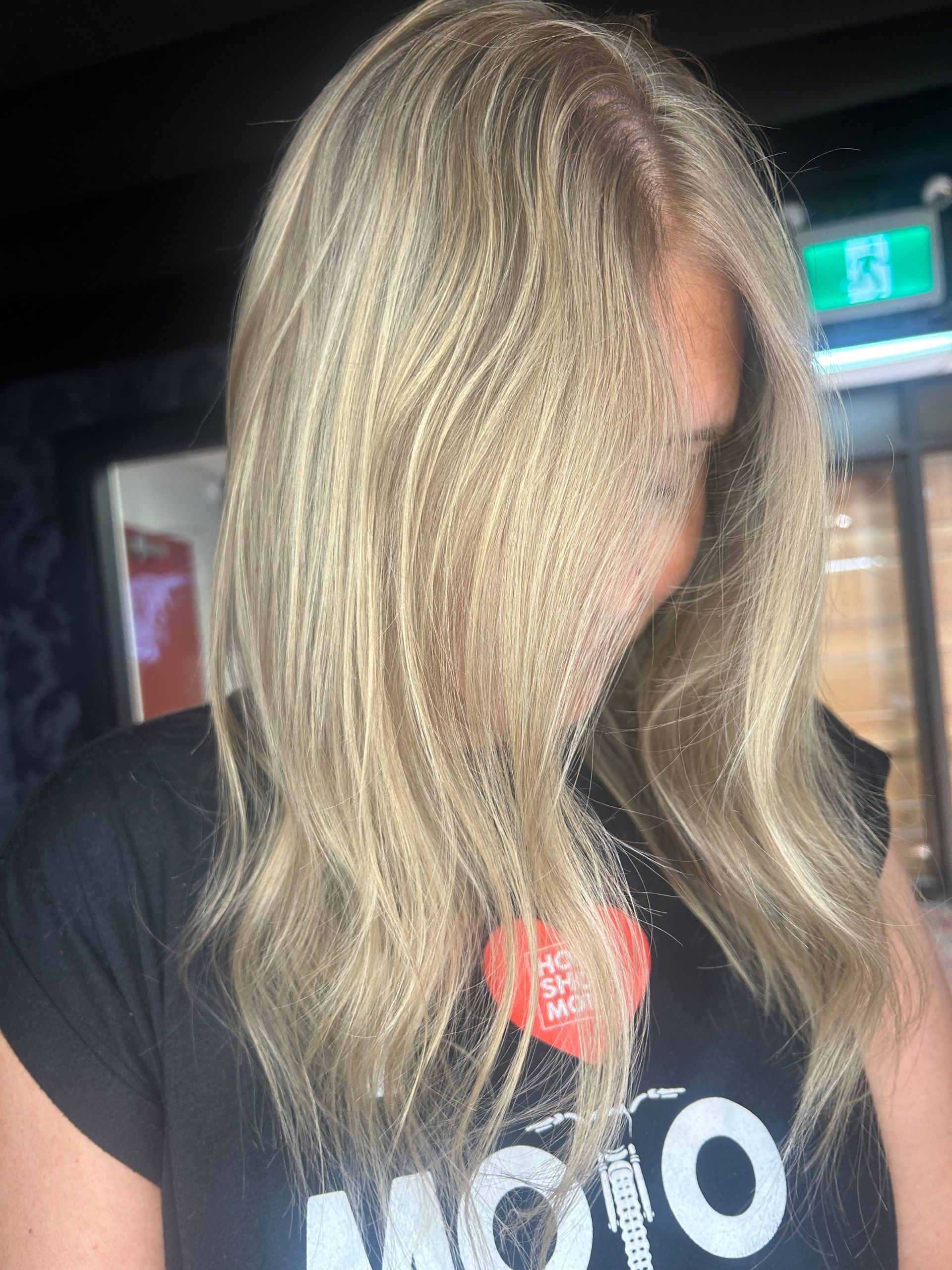 Person with long blonde hair wearing a black T-shirt with a red-and-white logo, indoors.