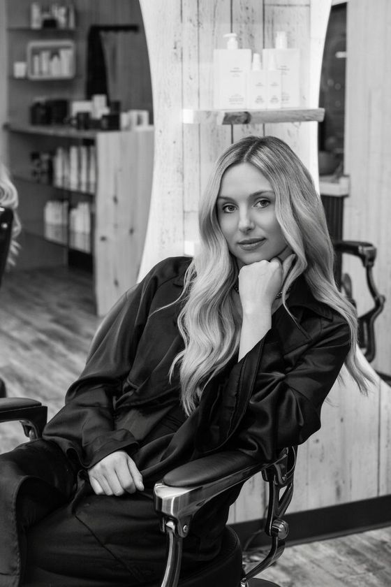 Woman sitting in a salon chair, resting her chin on her hand, with long hair and a relaxed expression.