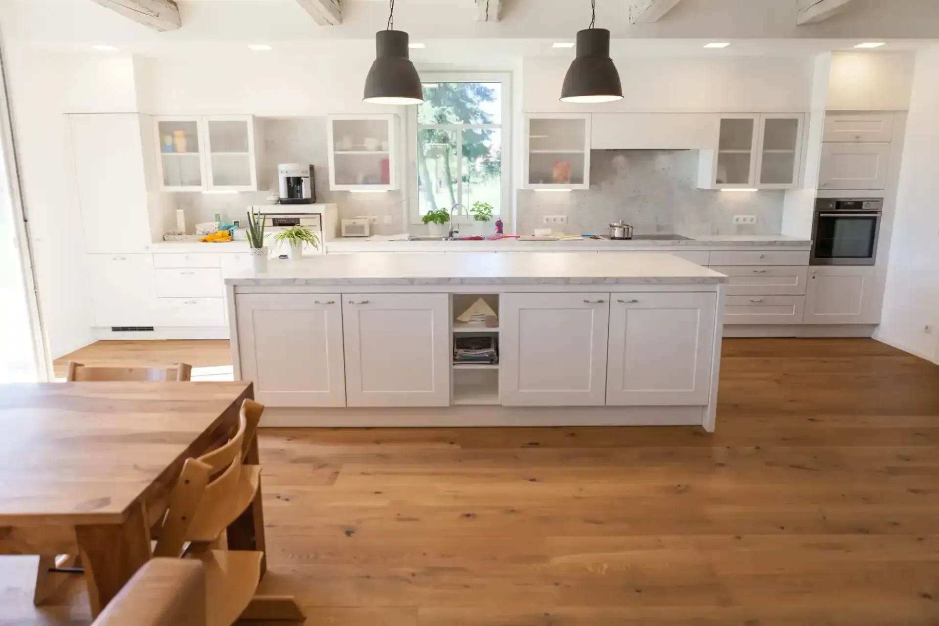 Geräumige weiße Küche mit Kochinsel, Holzboden und Beleuchtung. Im Vordergrund stehen ein Holztisch und Stühle.