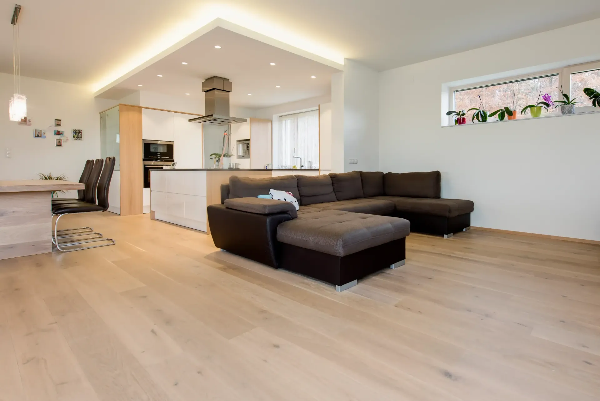 Lichtdurchfluteter offener Wohnbereich mit hellen Holzböden, einem dunkelbraunen Sofa und einer Küche im Hintergrund.