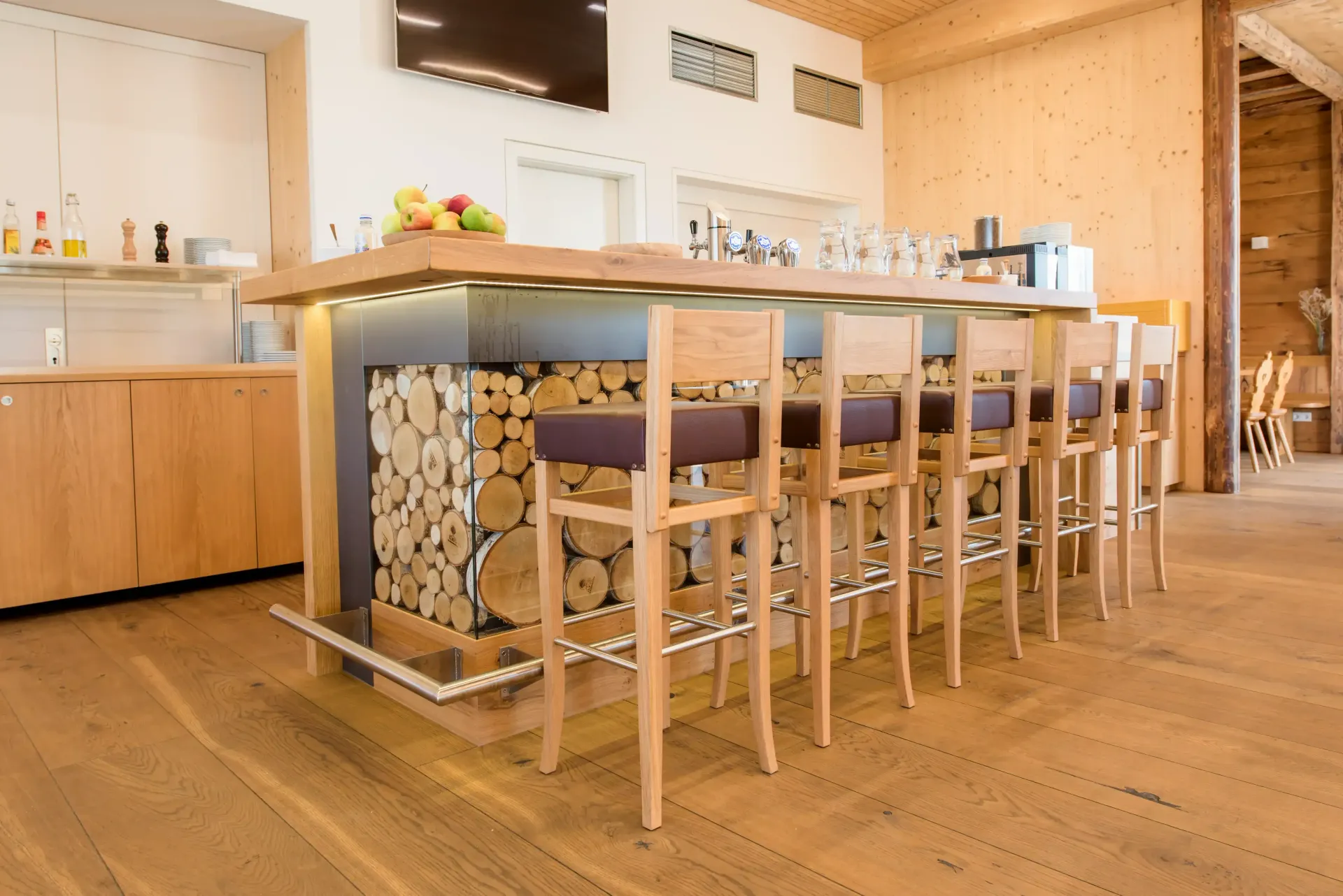 Bar mit Holzhockern und einer holzgetäfelten Front mit gestapelten Holzscheiten in einem Raum mit Holzboden.