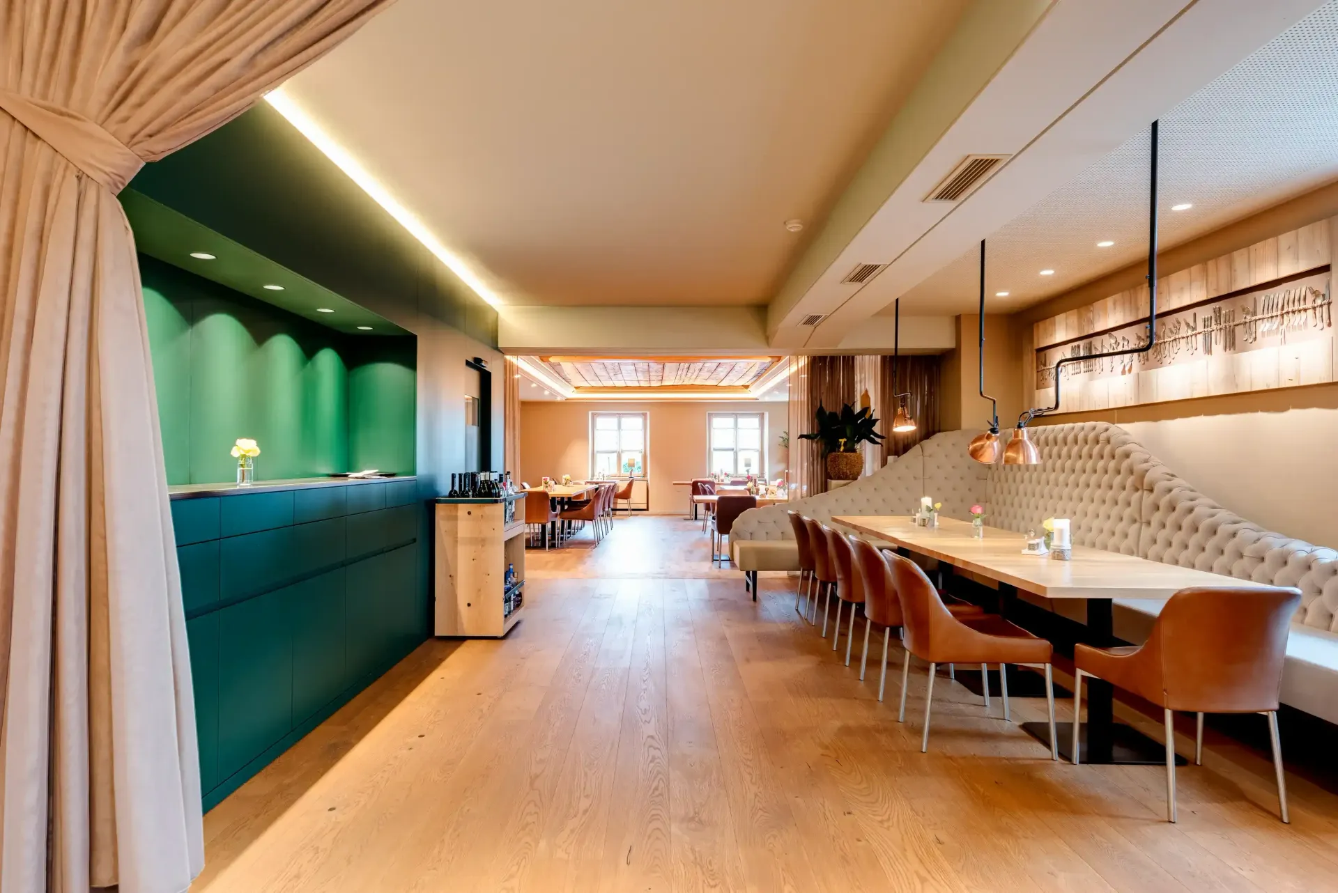 Inneneinrichtung des Restaurants: grüne und hellbraune Wände, langer Tisch mit braunen Stühlen, eingebaute Sitzecken, Barbereich.