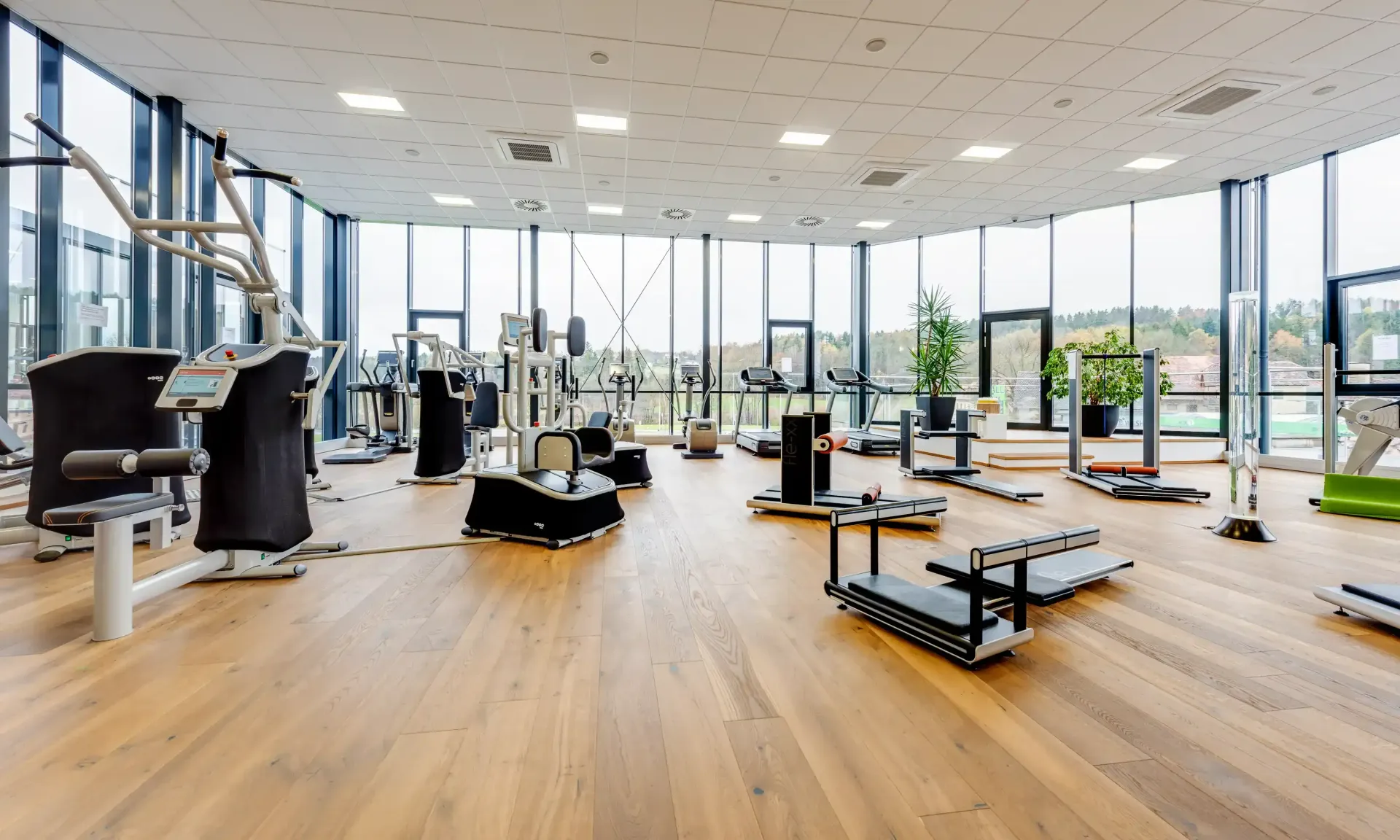 Fitnessstudio mit modernen Fitnessgeräten, großen Fenstern und Holzboden.