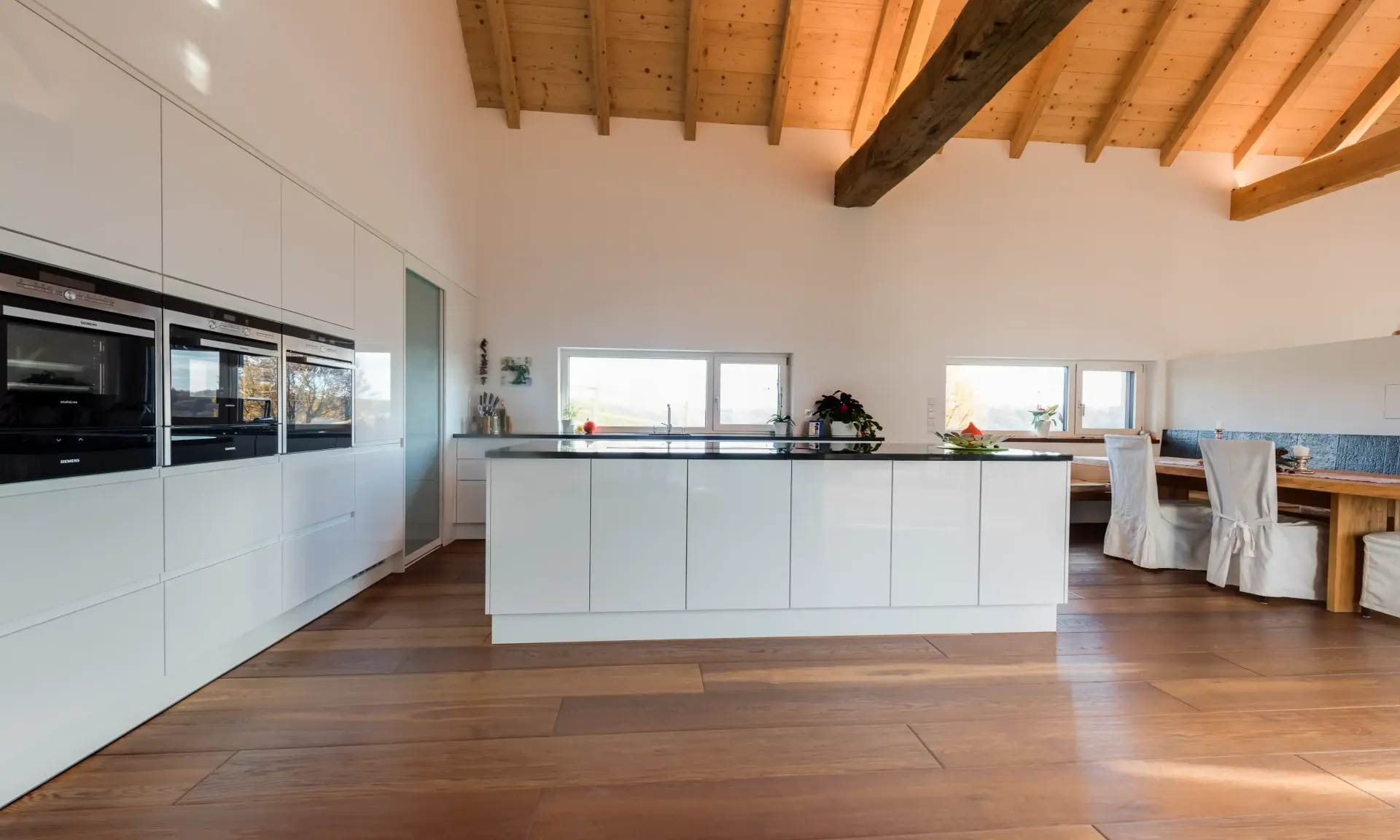 Moderne weiße Küche mit Holzböden und -decke, Kochinsel, Geräten und Essbereich.