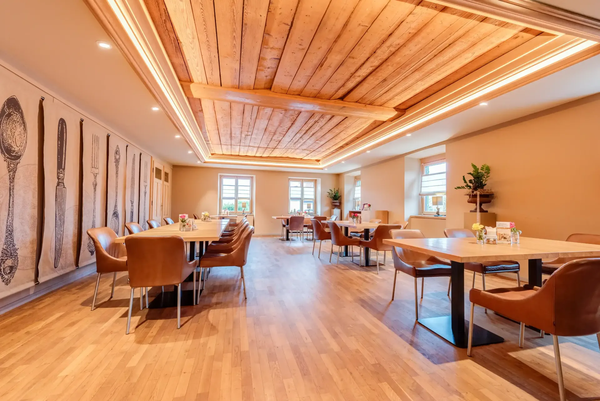 Restaurantinnenraum mit Holzdecke, Tischen, Stühlen und Küchenutensilienkunst an der Wand.