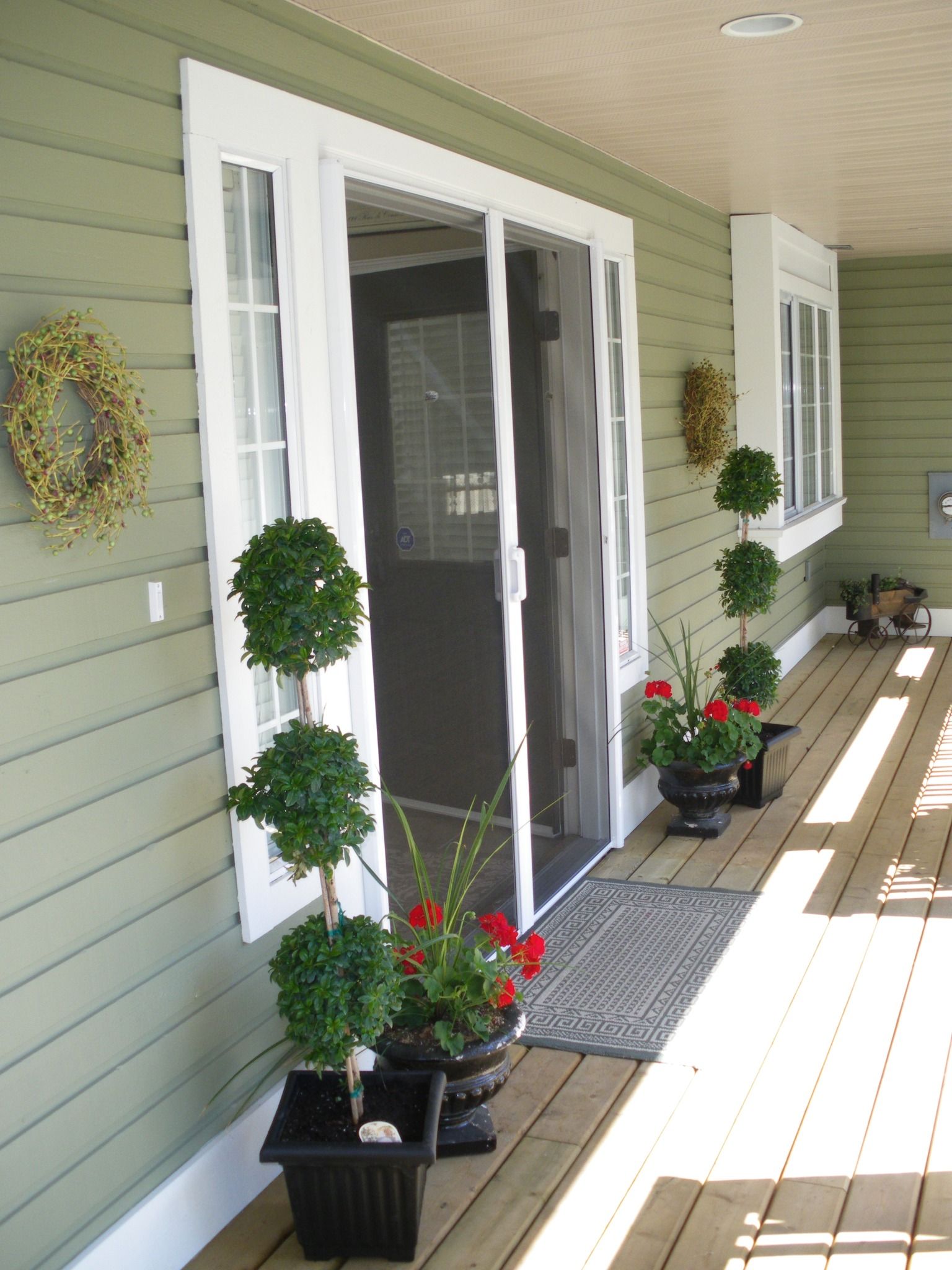 Porch with white sliding door, green siding, potted plants, and decorative wreaths in bright sunlight