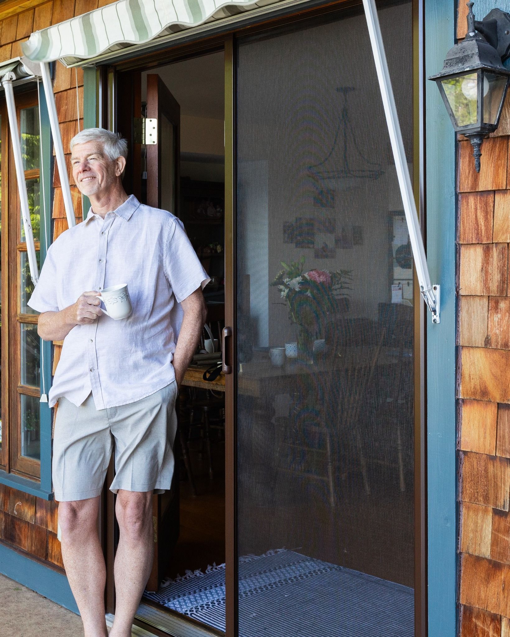 Man standing in a doorway on a porch, holding a mug beside a screen door.
