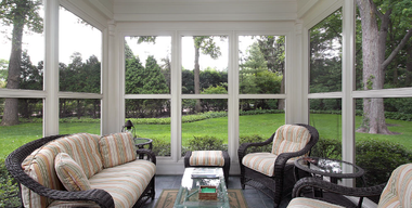 Porch with white sliding door, green siding, potted plants, and decorative wreaths in bright sunlight