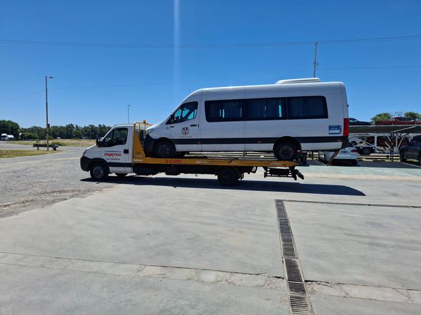 Una grúa carga un sedán plateado en una carretera costera bajo un cielo azul despejado.