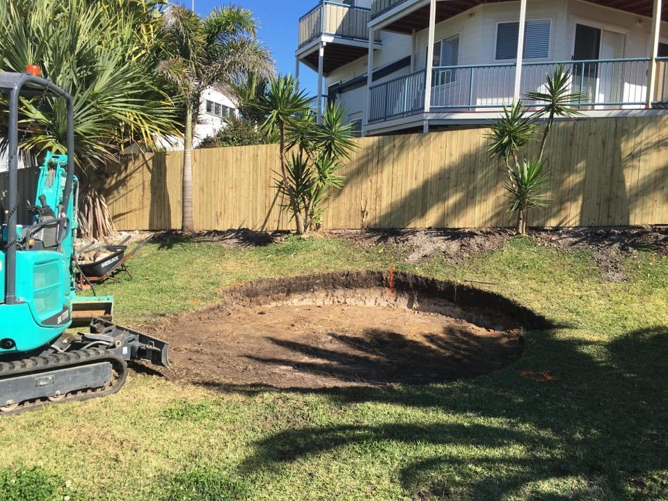A Small Excavator is Digging a Hole in the Grass in Front of a House — Caloundra Landscape Supplies in Caloundra, QLD