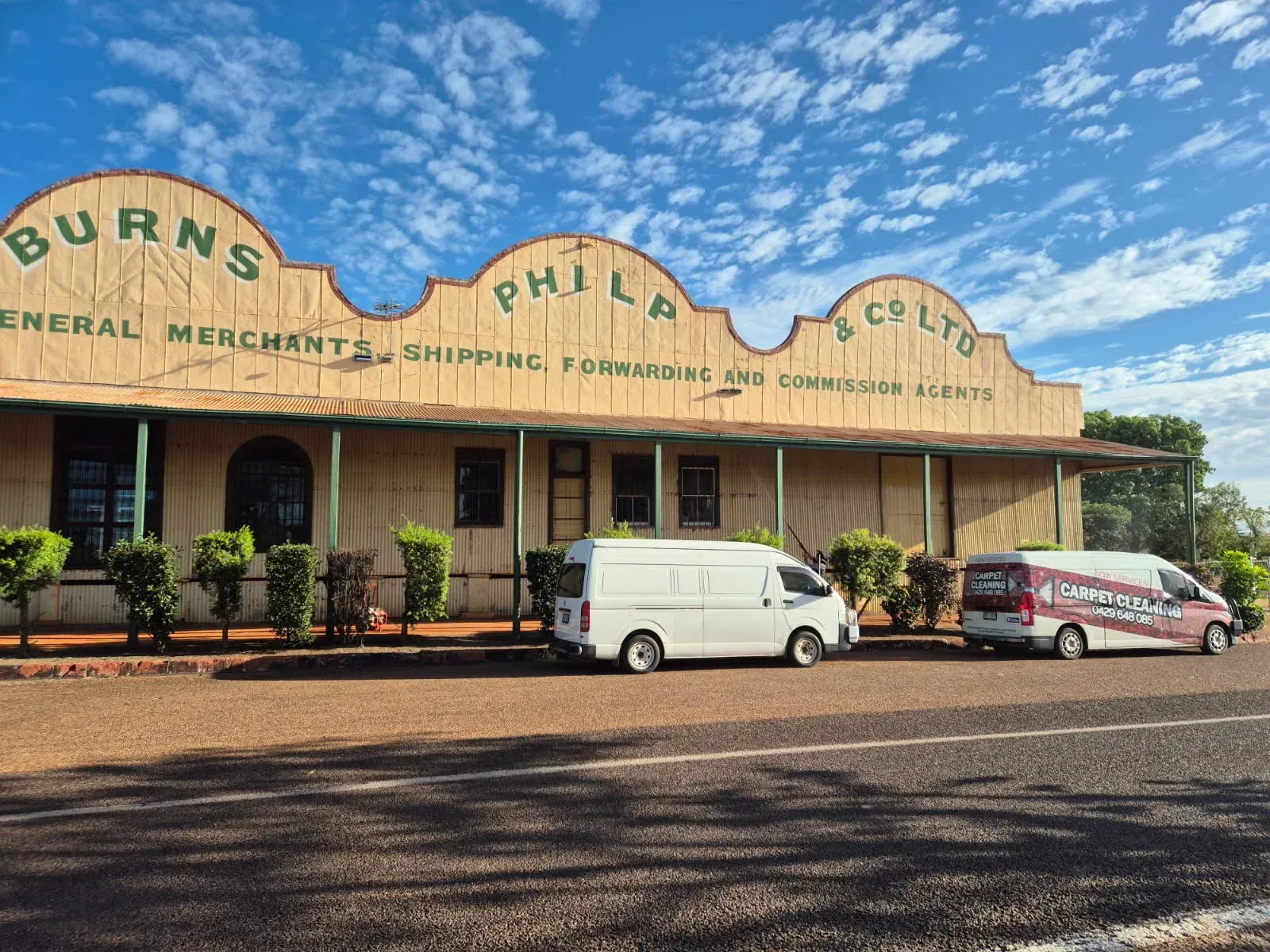 Small-town storefront with a tan facade, parked white van and red car under a bright blue sky. — NQW Services In Tully, QLD
