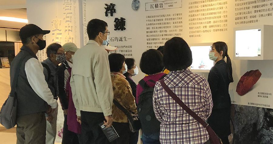 一位戴著口罩的解說員正在展覽館內向一群參觀者講解展品資訊。