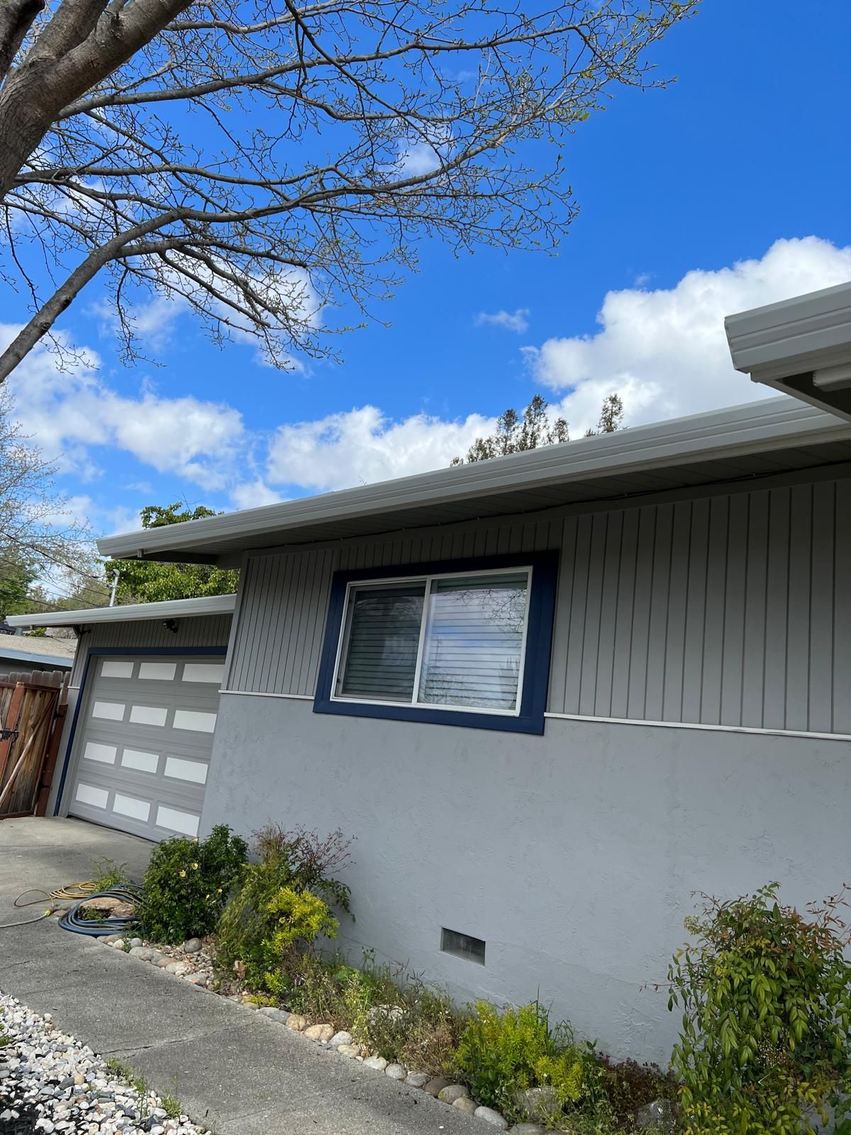 Suburban house exterior with gray siding, blue-trimmed windows, garage, and tree branches overhead