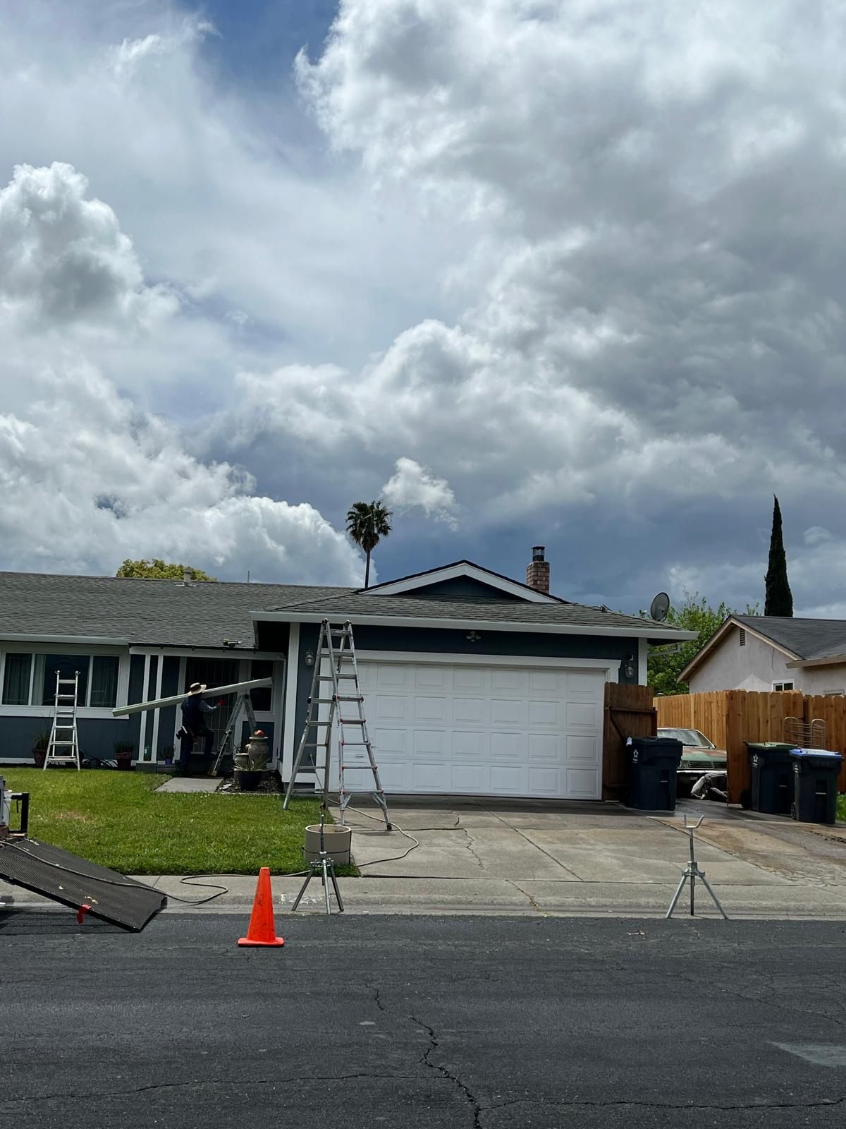 Single-story house with ladders and an orange cone in the driveway under a cloudy sky