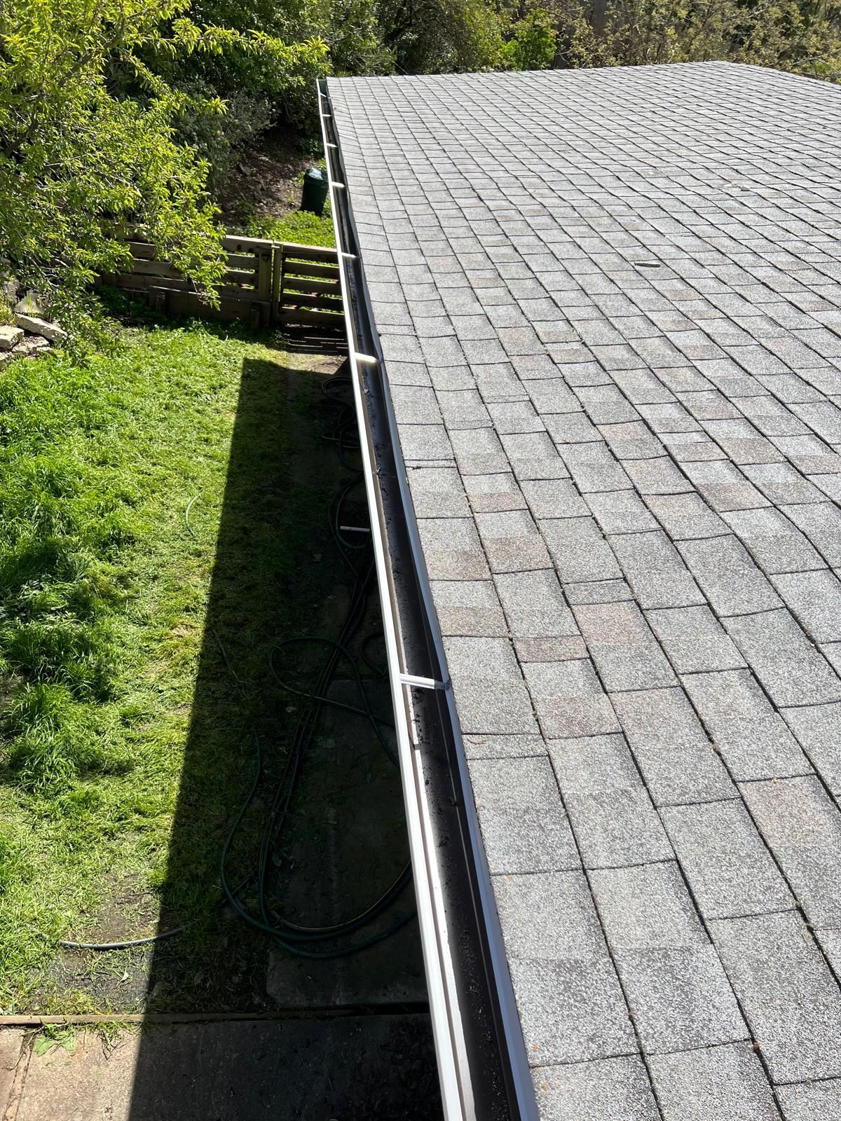 Roof with gray shingles beside a green yard and trees, viewed from above with a gutter along the edge