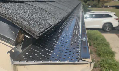 Close-up of a roof gutter with black perforated leaf guard beside dark shingles, with a white SUV in the background