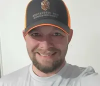 Smiling person in a black cap and light gray shirt against a plain white wall