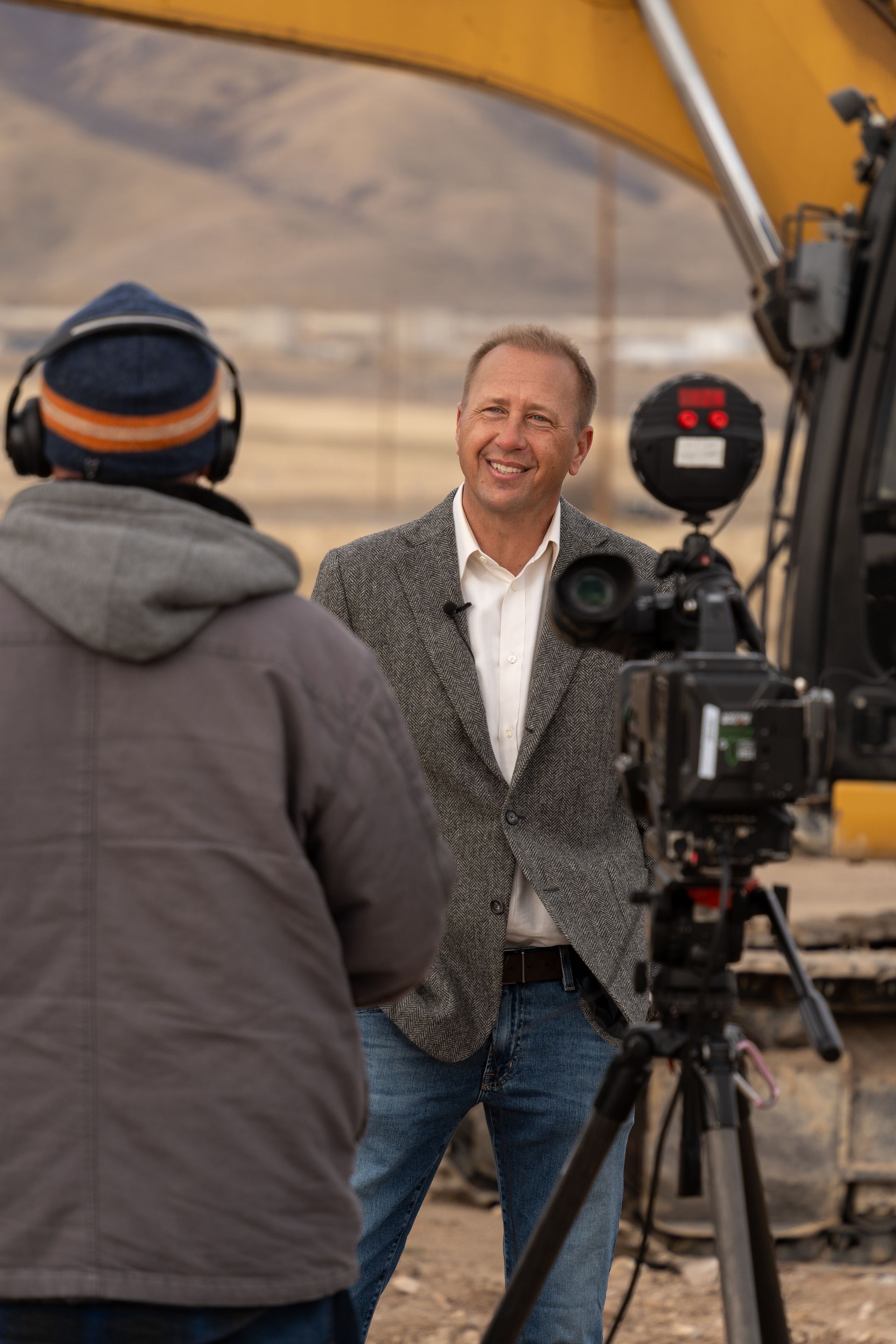 Rich Day standing at a build site with a reporter and news camera set up in front go him with an excavator in the background.