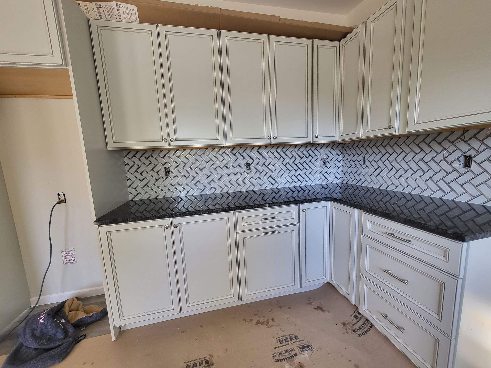 L-shaped kitchen with white cabinets, black countertop, and herringbone tile backsplash under construction