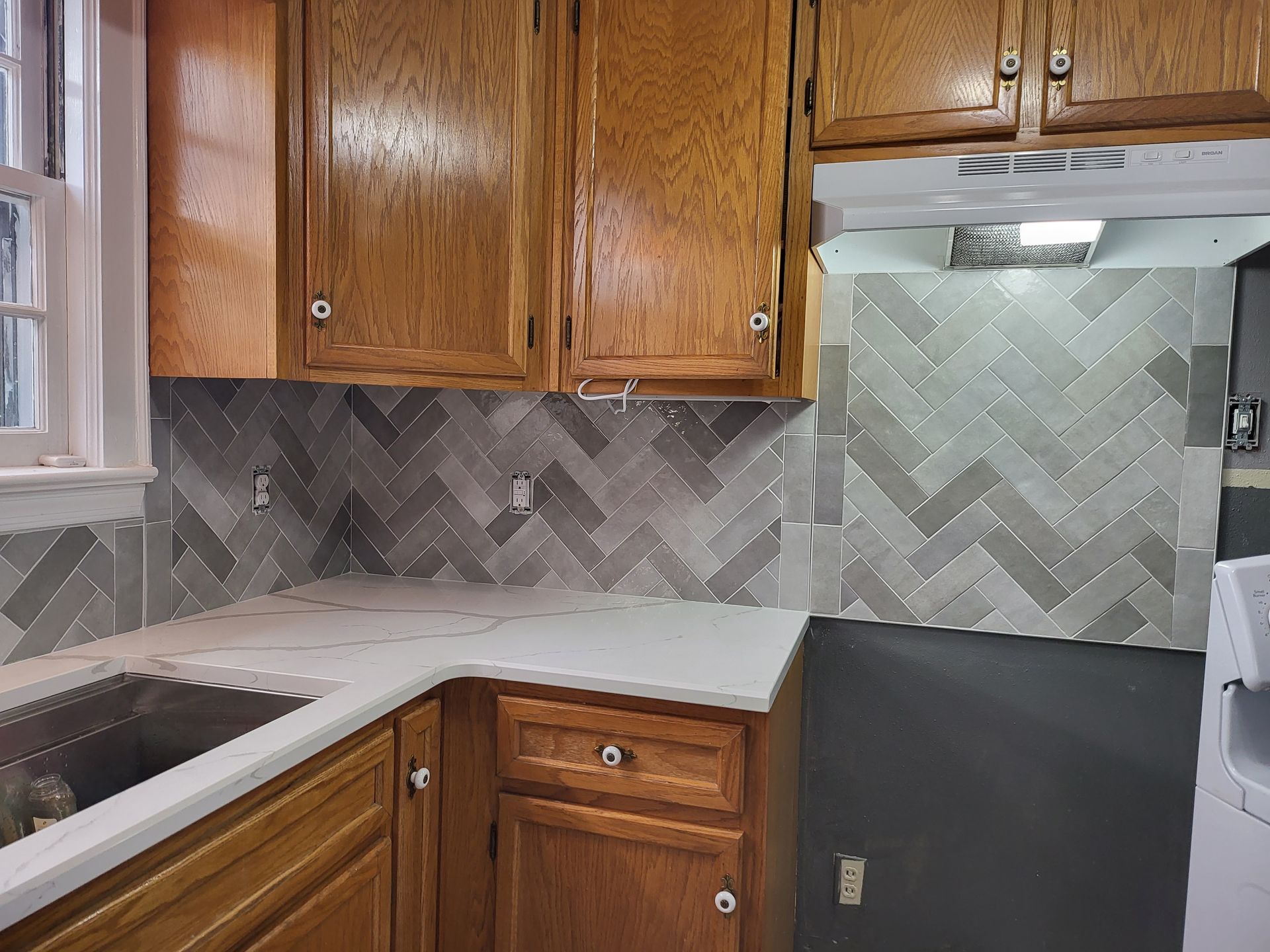 Kitchen with wood cabinets, white countertops, and gray herringbone tile backsplash, partially unfinished wall area.