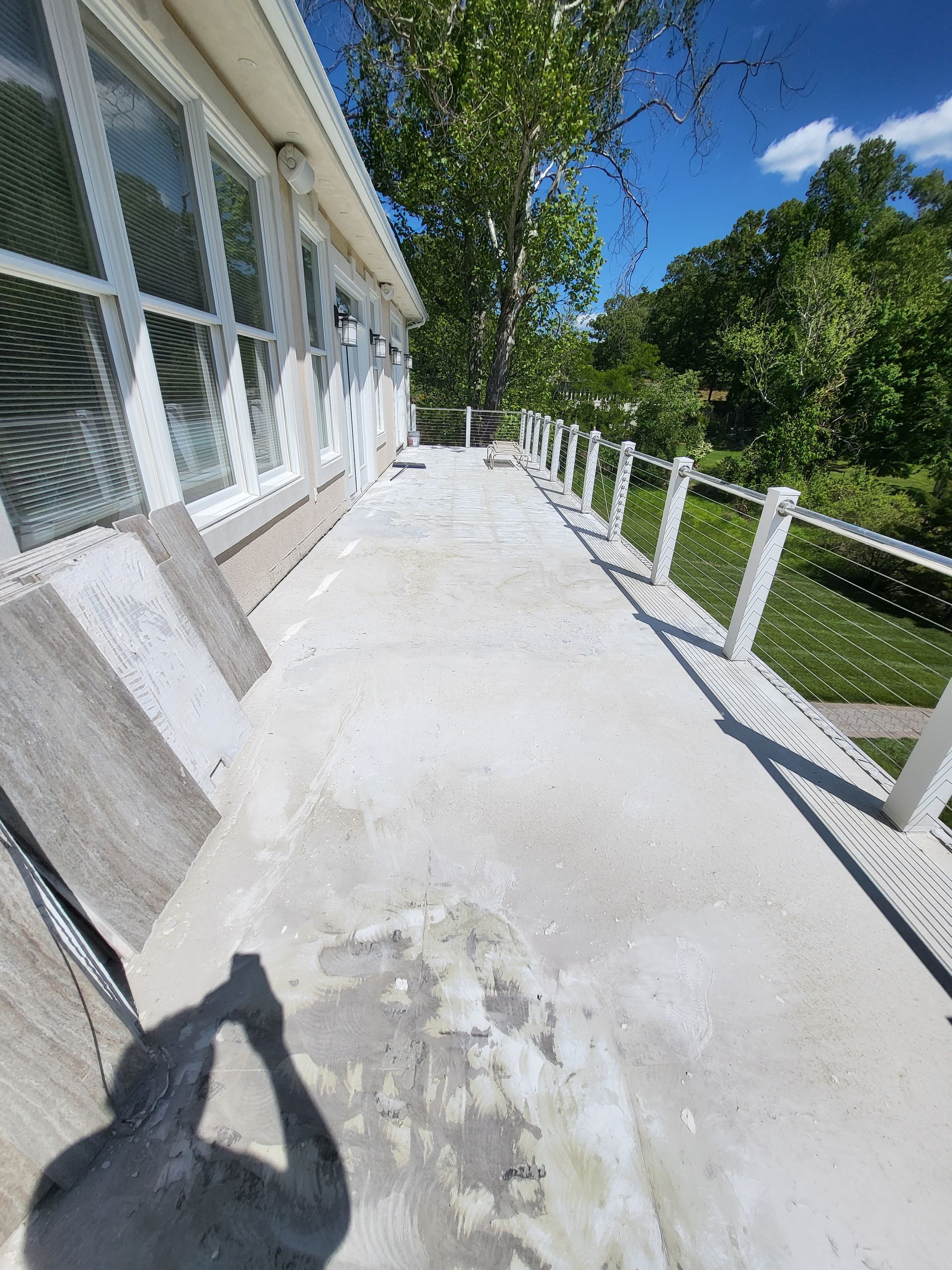 Concrete patio beside house with white railing, windows, and trees in bright sunlight