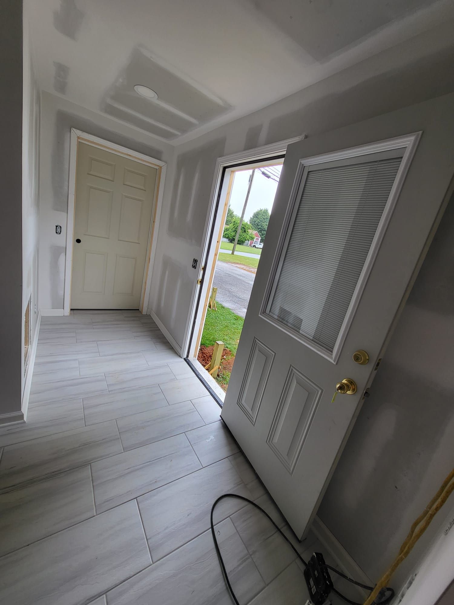 Front entry hallway with white door, tiled floor, and open front door showing a bright outdoor yard.