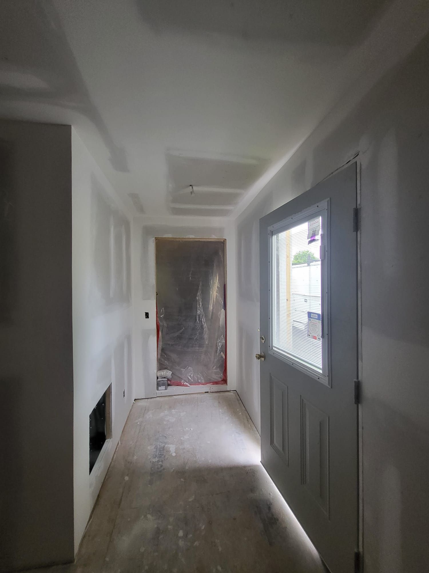 Narrow unfinished hallway with white walls, gray floor, and a windowed door on the right.