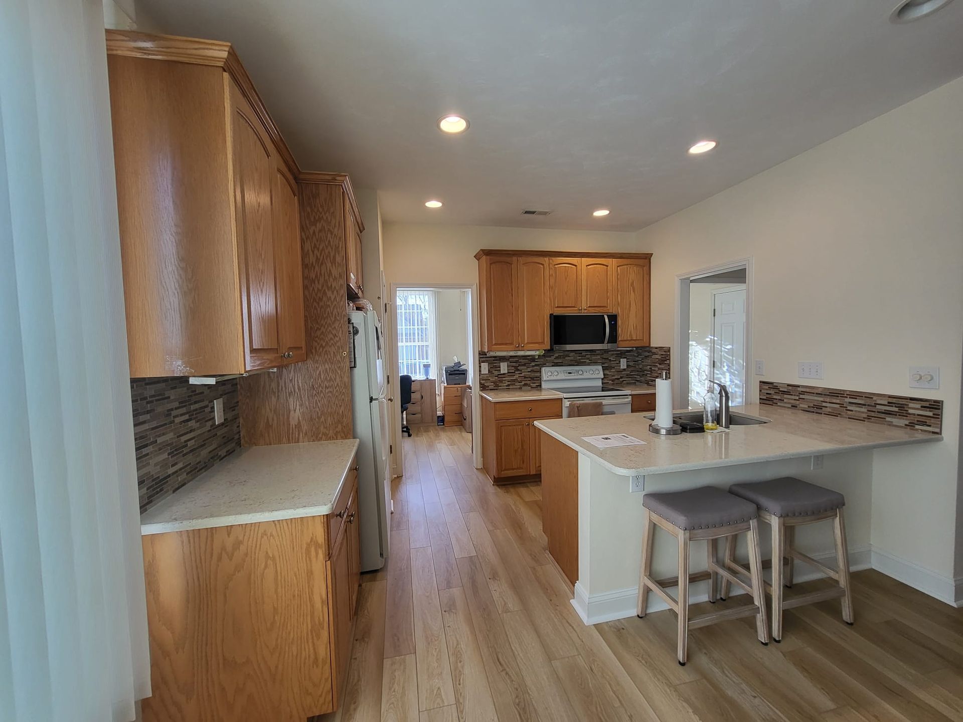 Empty kitchen with wood cabinets, light counters, breakfast bar, and two stools under recessed lighting.