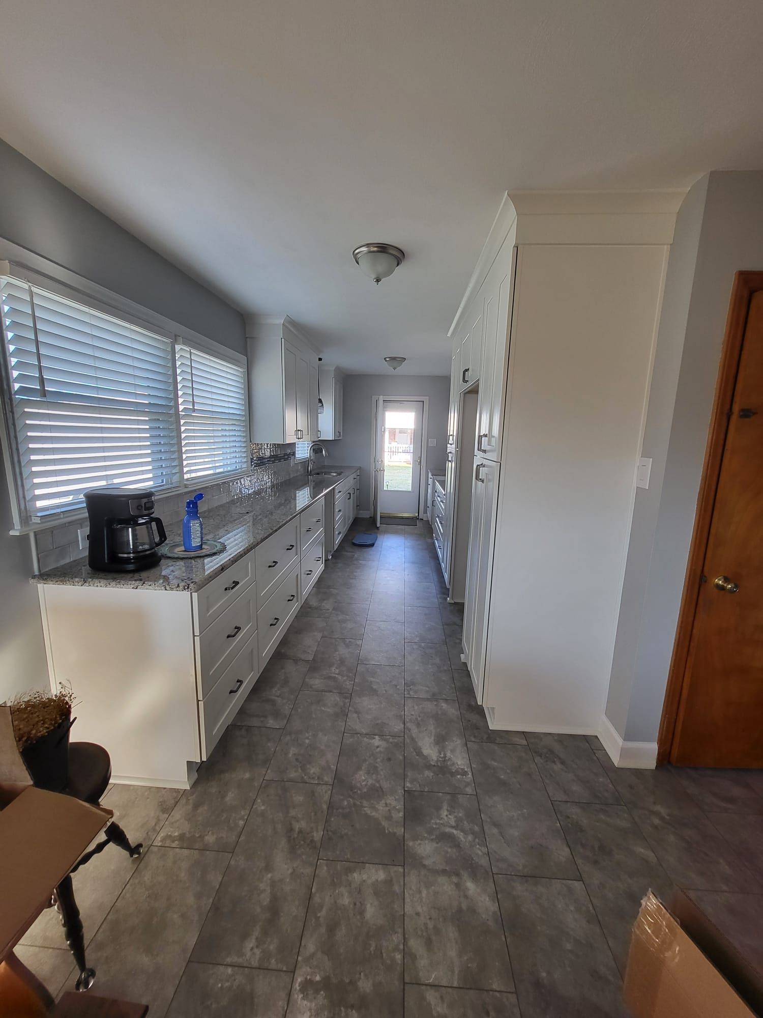 Narrow hallway kitchen with white cabinets, gray tile floor, and window blinds along the left wall