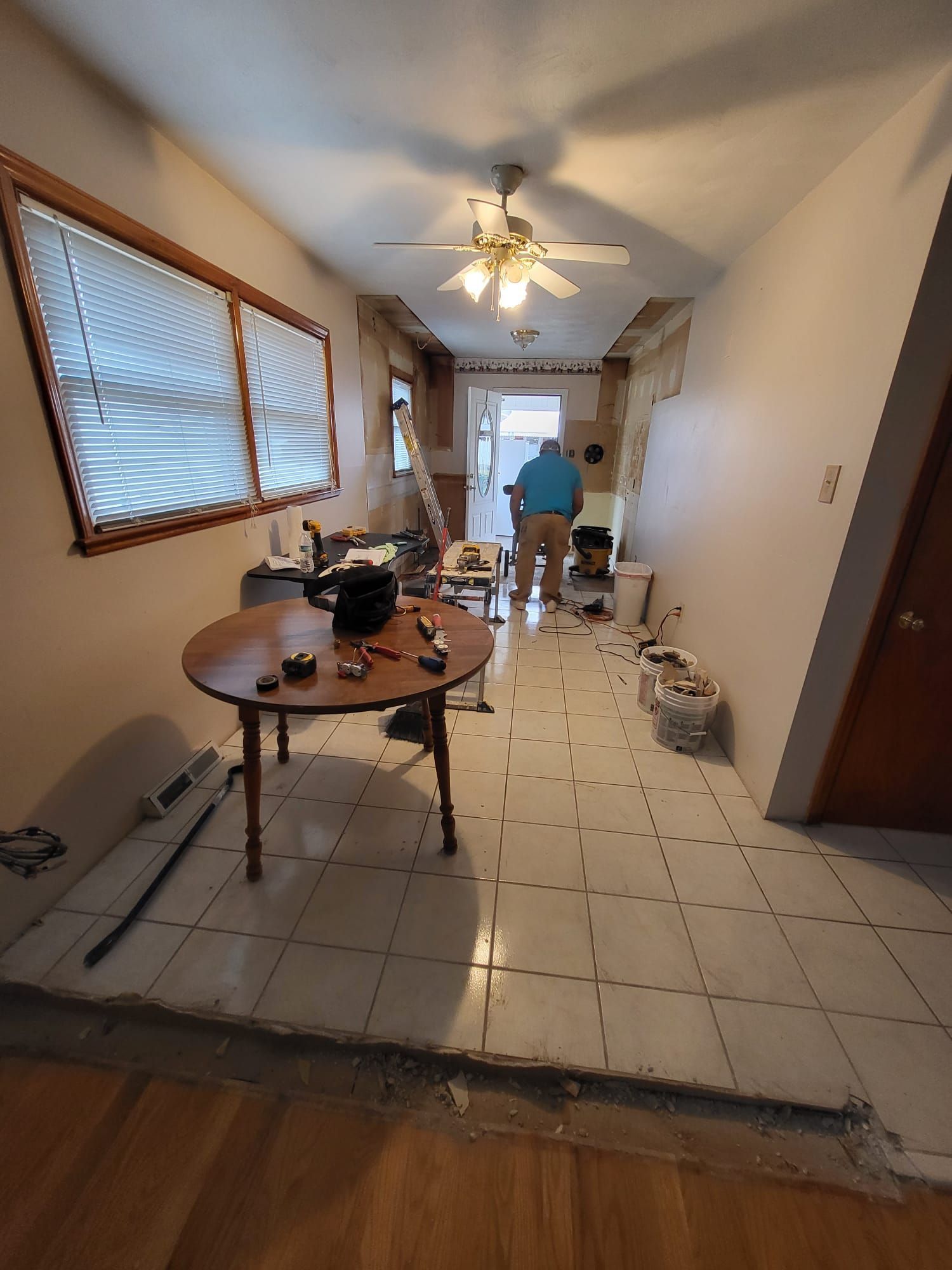 Narrow kitchen with white tile floor, ceiling fan, round table, and a person working near the back.