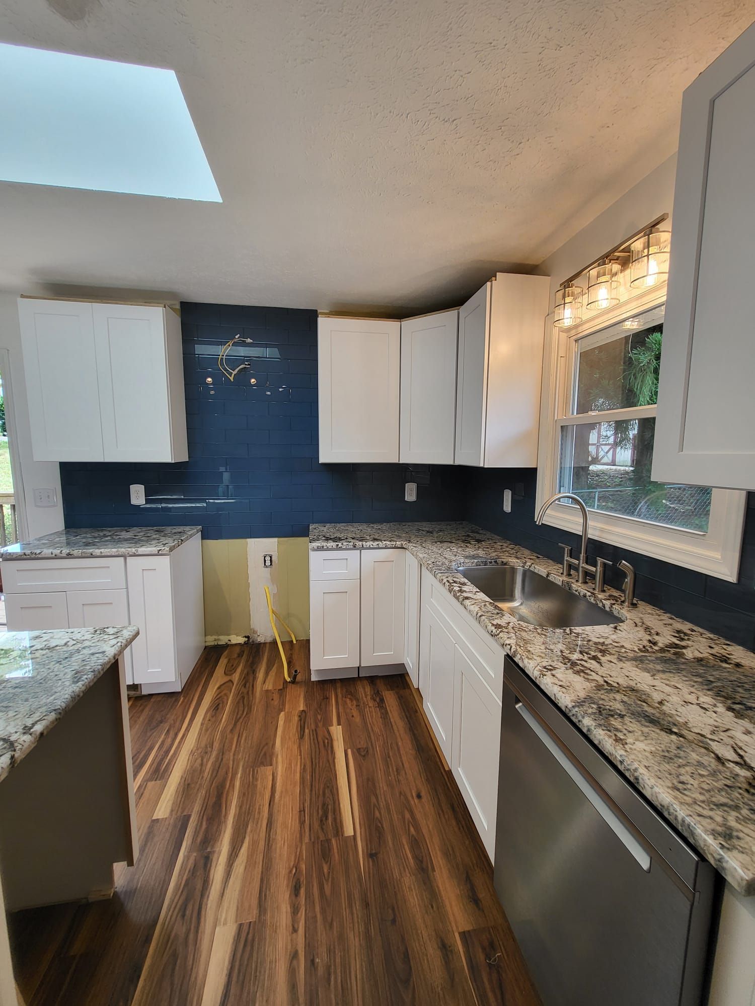 Small kitchen with white cabinets, dark blue backsplash, granite counters, wood floor, and a window over the sink.