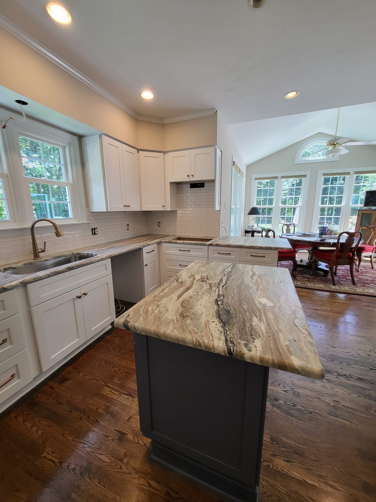 Bright kitchen with white cabinets, granite island, wood floors, and dining area near large windows.