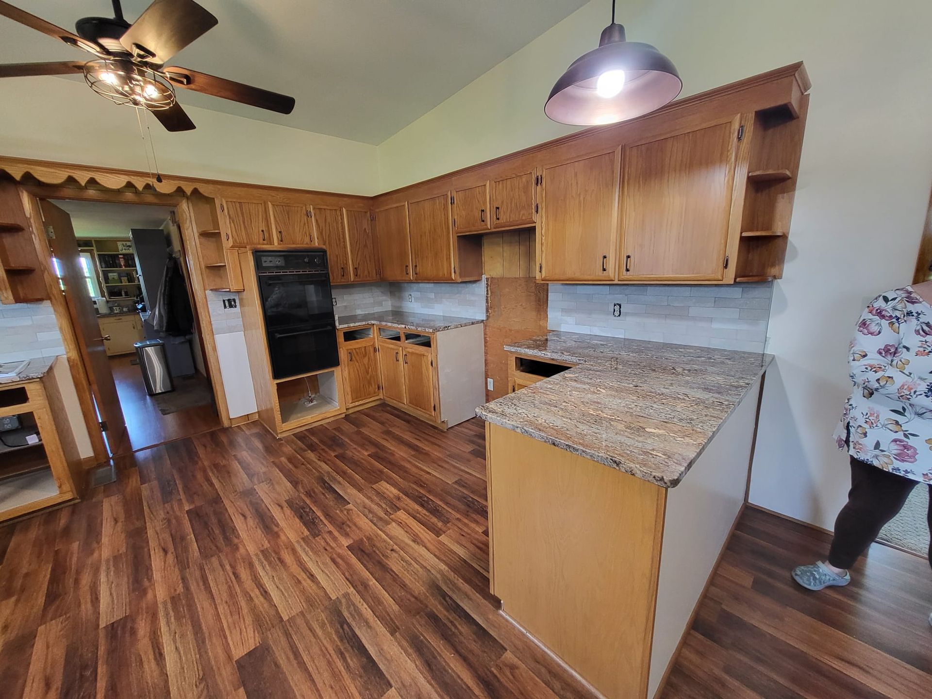 Empty kitchen with wood cabinets, granite island, ceiling fan, and hardwood floors; a person stands at right edge.