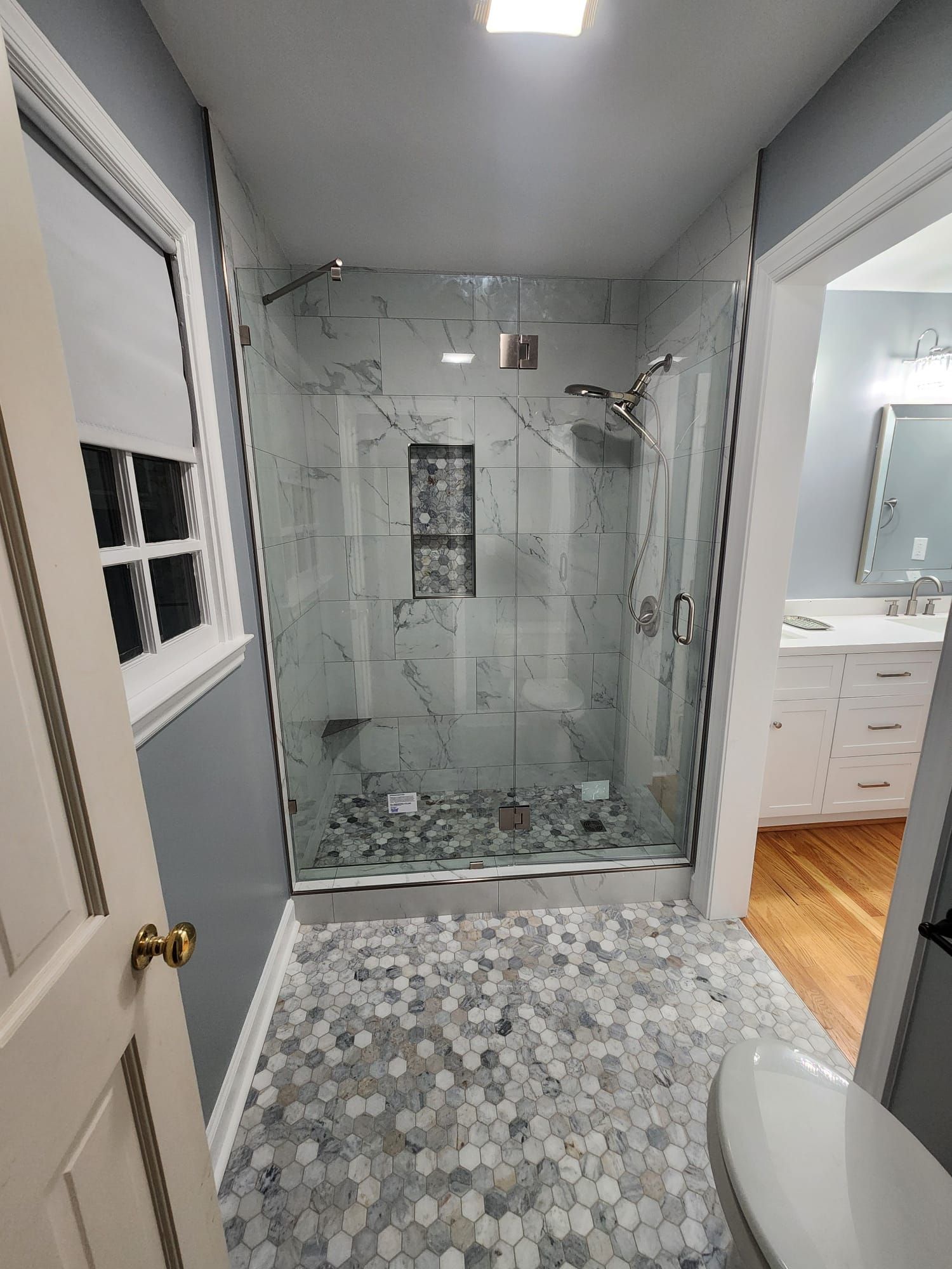 Small bathroom with glass shower, marble tile, and gray hexagon floor tiles leading to a vanity.